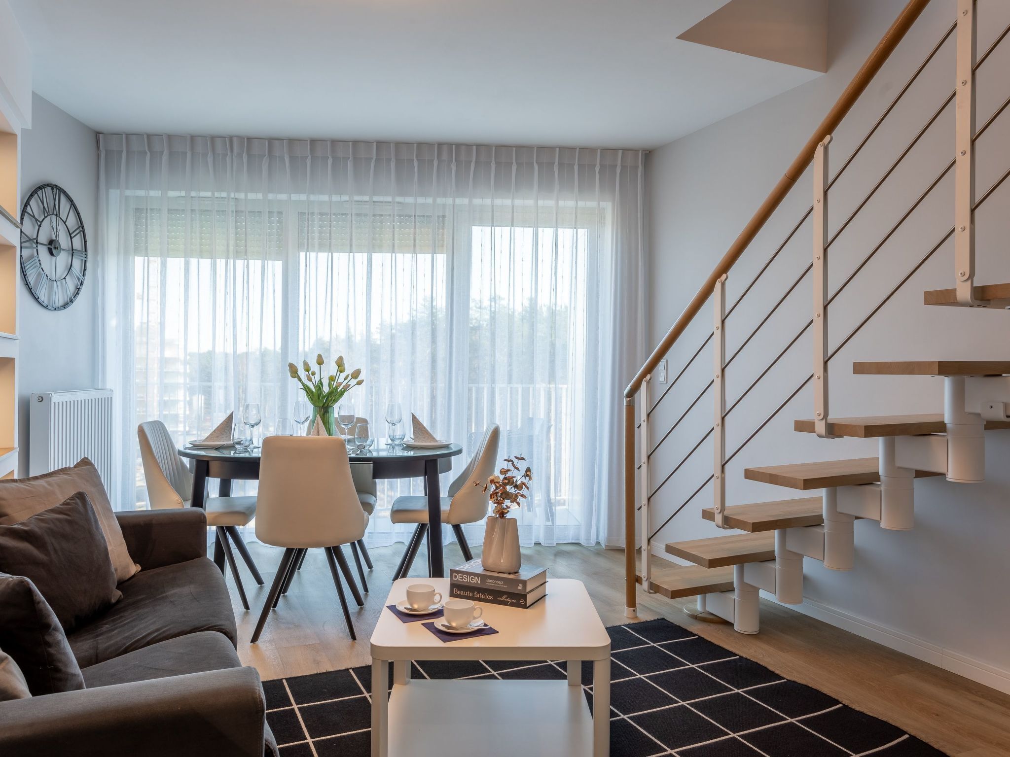 Una sala de estar moderna con una escalera, un comedor con una mesa preparada para dos personas y un ventanal con cortinas translúcidas.