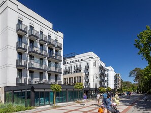 Ein modernes Gebäude mit mehreren Balkonen und großen Fenstern, umgeben von einer Fußgängerzone mit Bäumen und Bänken.