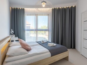 A modern bedroom with a bed, a bedside table, a window with curtains, and a view of buildings outside. - Urlaub auf Usedom - Ferienwohnung Tower 13 by Interhome (Swinoujscie)