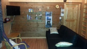 Une pièce chaleureuse avec un mur en bois, un canapé noir agrémenté d’un coussin, un fauteuil en osier et une bibliothèque contenant divers objets.