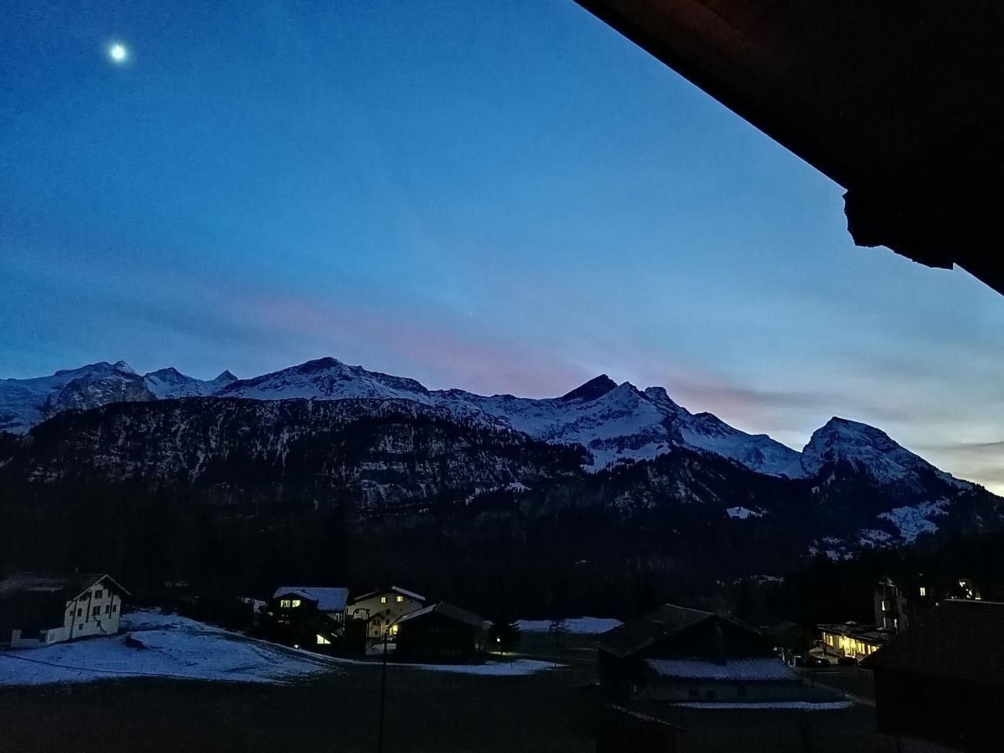 A mountain landscape at dusk with snow-covered peaks and a few buildings in the foreground.