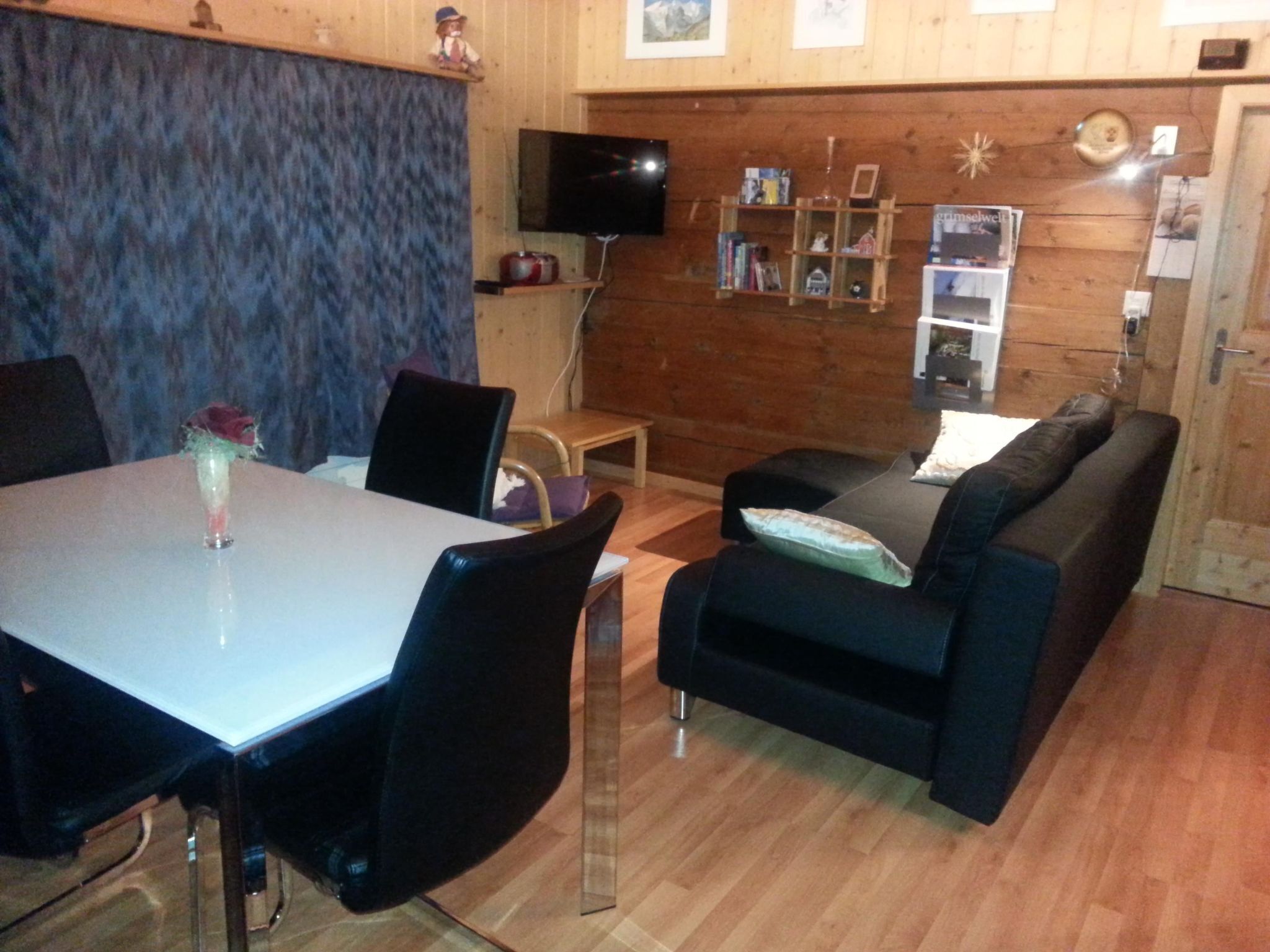 En hyggelig stue med spisebord, sorte stole, en sort sofa og et fjernsyn monteret på væggen.