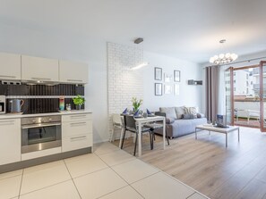 Eine moderne Küche mit weißen Schränken, einer schwarzen Spritzschutzwand und einem Essbereich mit Tisch und Stühlen. Im Wohnbereich befindet sich eine Couch und ein Couchtisch, und eine Glastür führt auf einen Balkon.