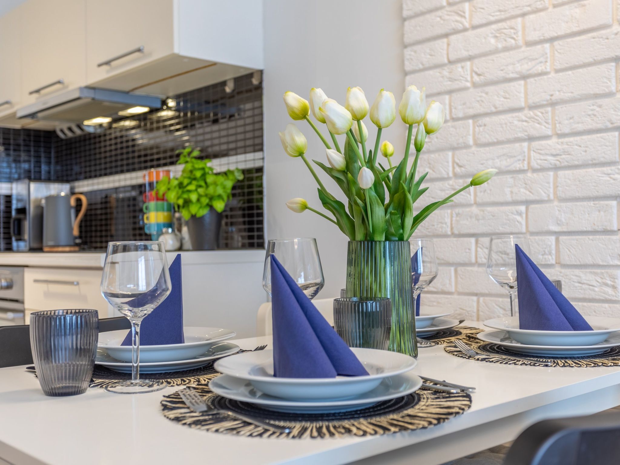 Une cuisine moderne avec des murs en briques blanches, un dosseret de carreaux noirs et un coin repas avec une table blanche mise pour deux personnes.