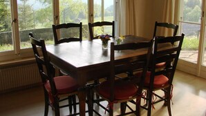 Une table à manger avec six chaises, un vase rempli de fleurs et une vue sur les arbres et l’herbe, visible à travers de grandes fenêtres.