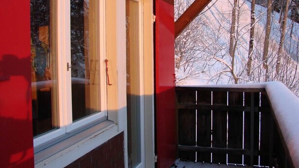 Une fenêtre avec une porte rouge, une balustrade en bois et un paysage enneigé en arrière-plan.