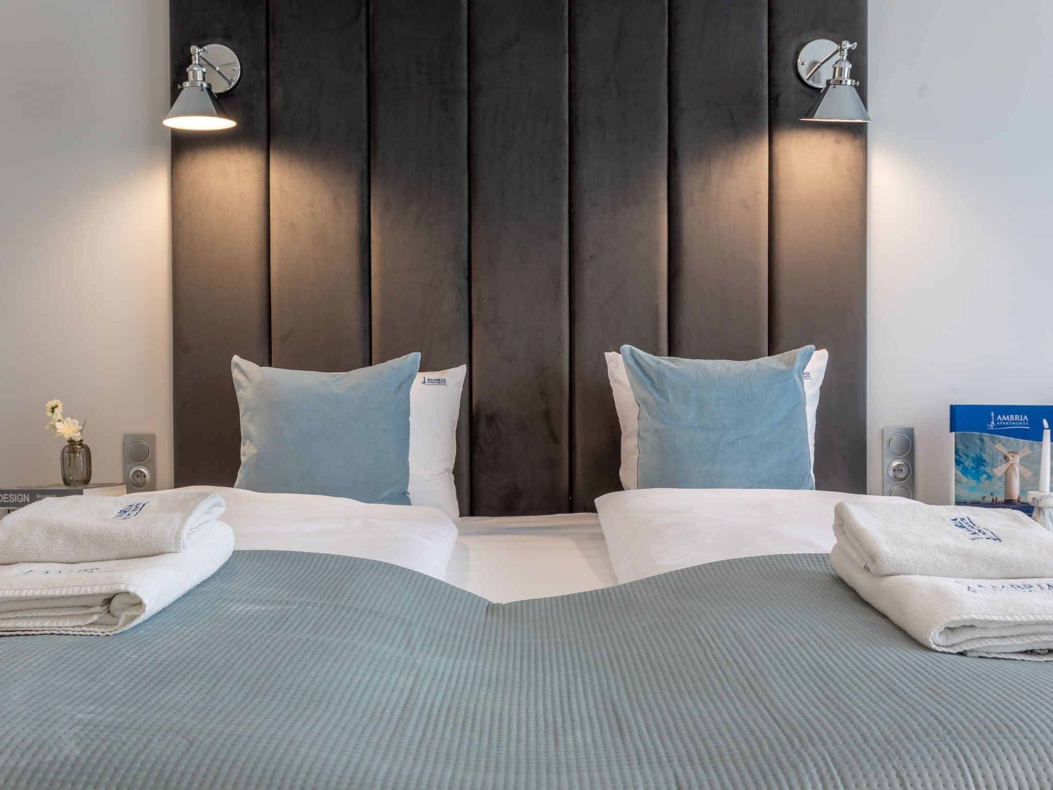 Habitación de hotel con cabecera de madera oscura, dos almohadas, una toalla doblada y un libro en la mesita de noche.