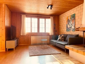 Ein gemütliches Blockhauszimmer mit Holzwänden, einer Couch, einem Fernseher und einem großen Fenster mit Vorhängen.