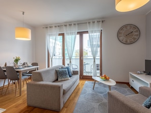 Ein modernes Wohnzimmer mit einer Couch, einem Esstisch, Stühlen, einem kleinen Tisch mit Obst und einem großen Fenster mit Vorhängen.