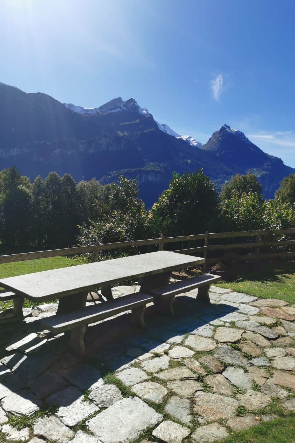 Un tavolo da picnic con panche su una zona pavimentata in pietra, circondato da una recinzione in legno, con un paesaggio montuoso sullo sfondo.