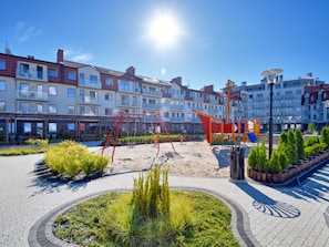 A playground with swings and climbing structures, surrounded by a paved area with greenery and a row of multi-story buildings in the background. - Ferienappartement del Sol auf Usedom by Interhome (Swinoujscie)