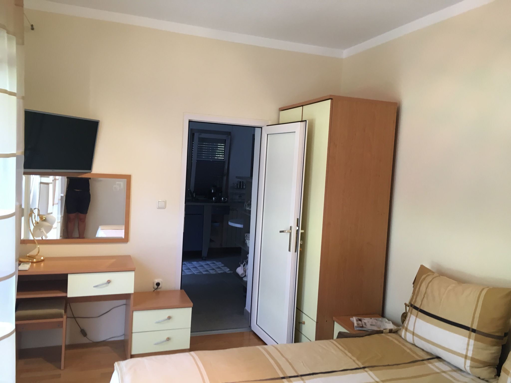 A bedroom with a bed, a wardrobe, a desk with a chair, a mirror, and a television on the wall.