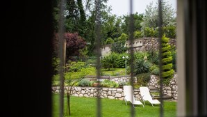 A view of a garden with a stone wall, a wooden bench, and a white chair through a window with vertical blinds. - mit Terrasse by Interhome (Opatija/Rukavac)