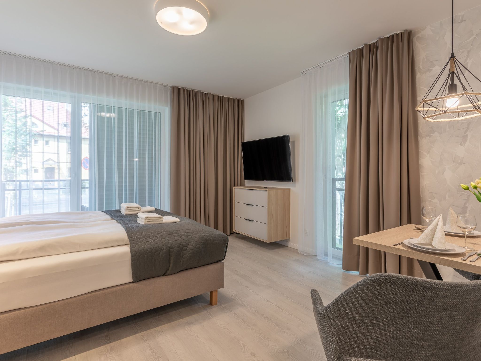 A modern hotel room with a large bed, a TV, a desk with a chair, and a dining area.