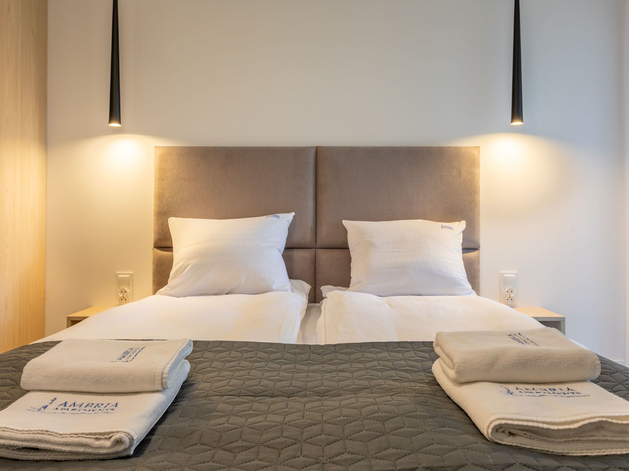 Una cama doble con colcha gris acolchada, dos almohadas blancas y dos toallas dobladas con texto.