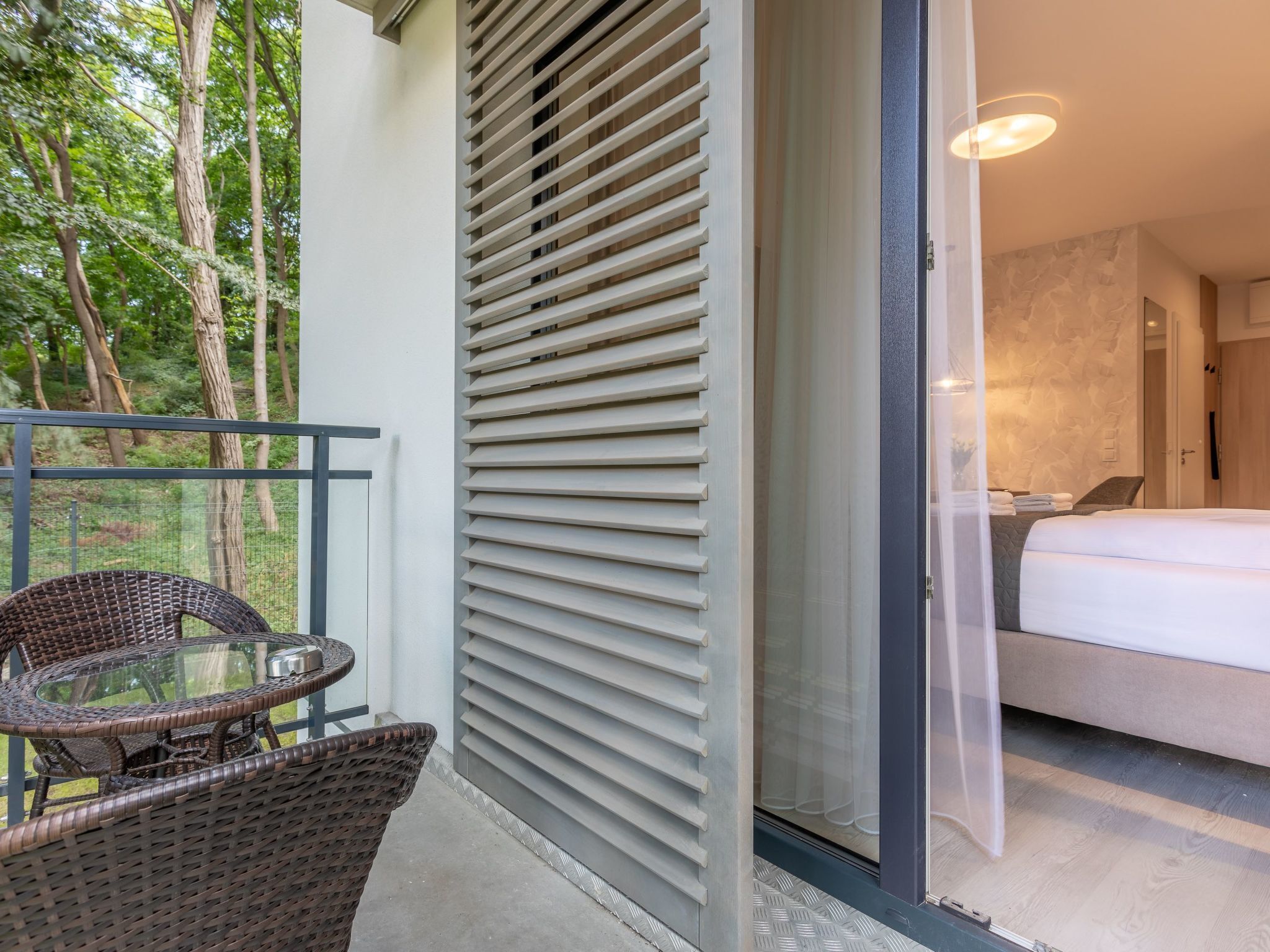 Un balcon doté de meubles en osier, une porte-fenêtre coulissante donnant sur une chambre à coucher avec un lit et une table de chevet.