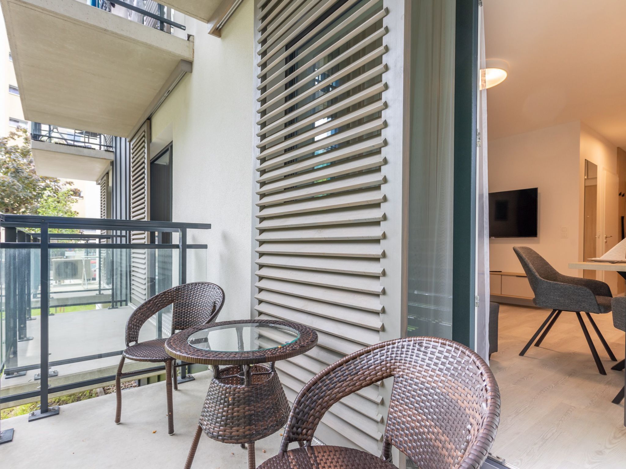 Un balcón con sillas de mimbre, una mesa de vidrio y una puerta corrediza de cristal, además de un televisor en la habitación.