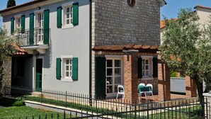 Una casa de dos pisos con balcón, sección de ladrillo a la derecha y un patio cercado.