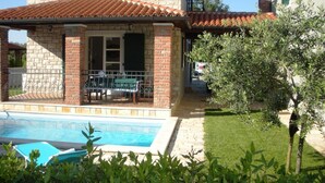 Una casa de dos pisos con piscina, un patio con mobiliario exterior y un jardín con olivos.