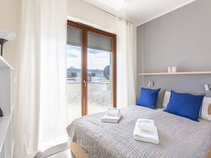 A hotel room with a bed, a window with blinds, a ceiling fan, and a view of the cityscape. - Sky mit Balkon am Meer by Interhome (Swinoujscie)