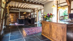Um quarto amplo com uma grande escrivaninha de madeira, um computador e um vaso de flores. Há uma área de refeições com cadeiras e uma mesa, e uma cozinha com armários e pia.