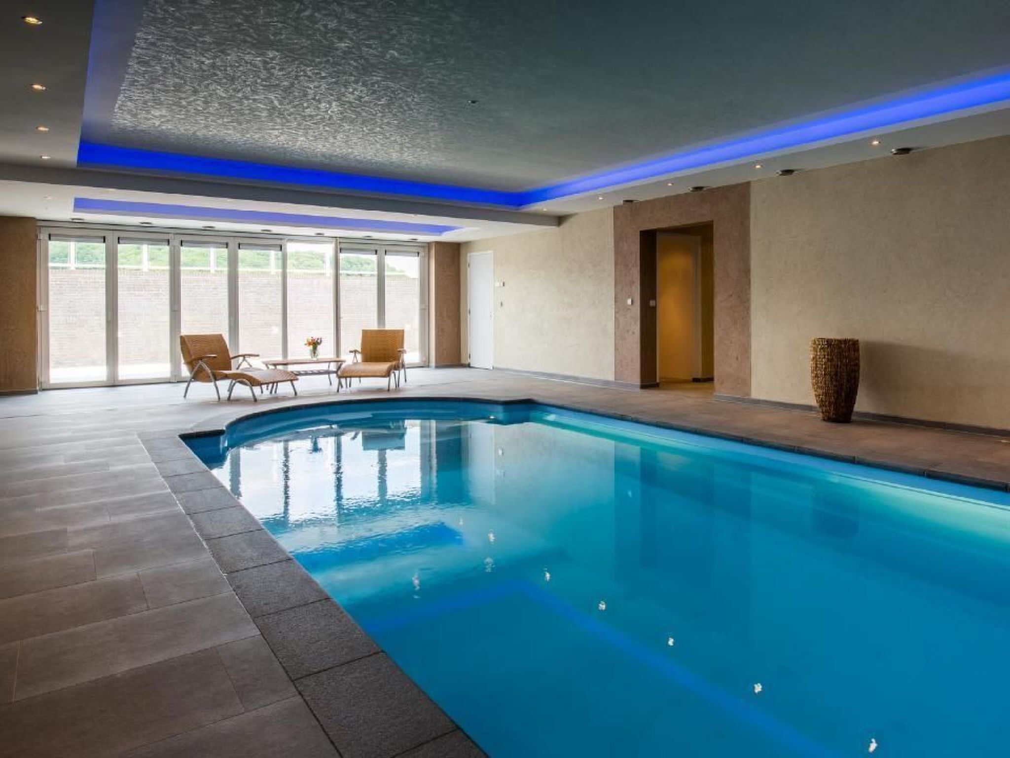 A swimming pool with a tiled floor, a few lounge chairs, and a door in the background.