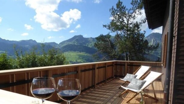 Una terraza de madera con dos sillones de descanso y dos copas de vino, con vistas a un paisaje montañoso.
