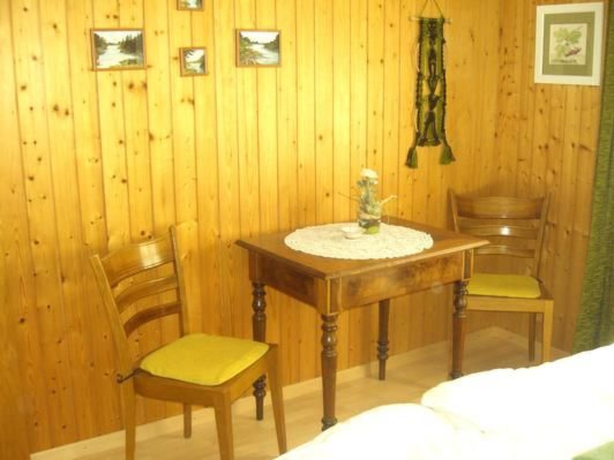 Une pièce en bois avec un lit, deux chaises, une table sur laquelle se trouve un vase, et des tableaux encadrés accrochés au mur.