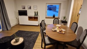 A modern living room with a wooden dining table, comfortable chairs, a flat-screen TV, a white entertainment unit, and a window with a view of a snowy landscape.