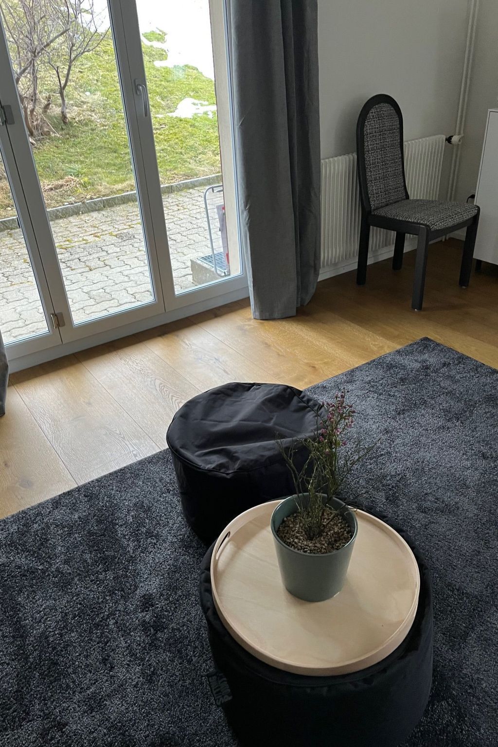 Ett rum med ett stort fönster, en stol, en svart pall och en krukväxt på ett runt bord.