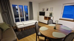 A compact living space with a sofa, a small bed, a dining table with chairs, a TV, and a window with a view.