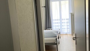 A narrow hallway leading to a bedroom with a bed, a small stool, and a window with curtains.