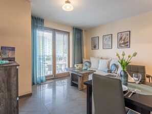 A modern living room with a sofa, coffee table, and dining table. There are large windows with blinds, a ceiling light, and framed artwork on the wall. - Ferienappartement Aurora auf der Insel Usedom by Interhome (Swinoujscie)
