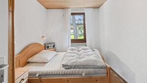 Quarto com cama de madeira, mesa de cabeceira, rádio e janela com vista para o verde.
