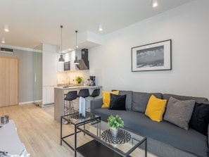 Ein modernes Wohnzimmer mit grauem Sofa, schwarzen und gelben Kissen, einem Glastisch und einer Küche im Hintergrund.