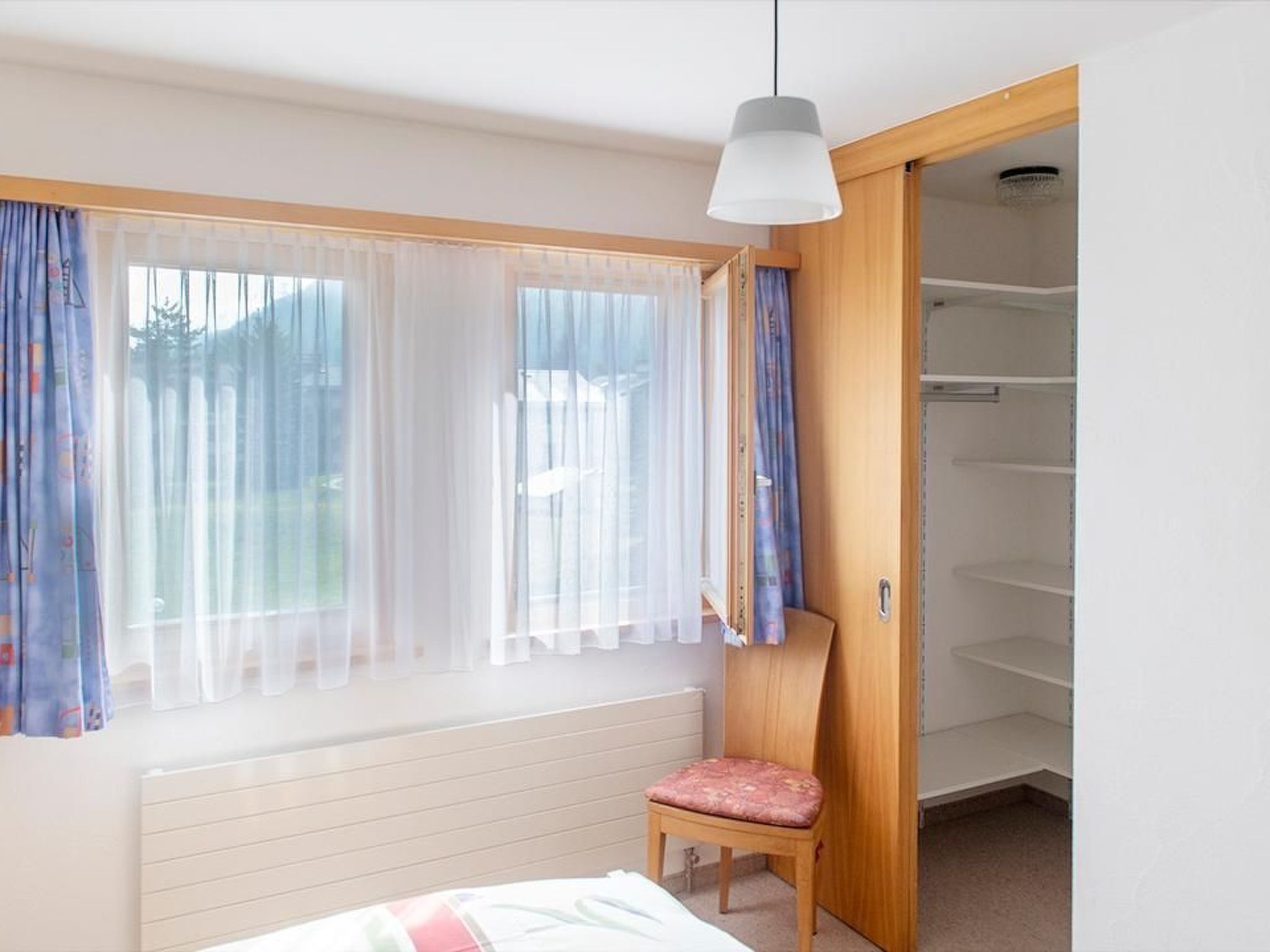 A bedroom with a bed, a chair, a wardrobe, and a window with curtains.