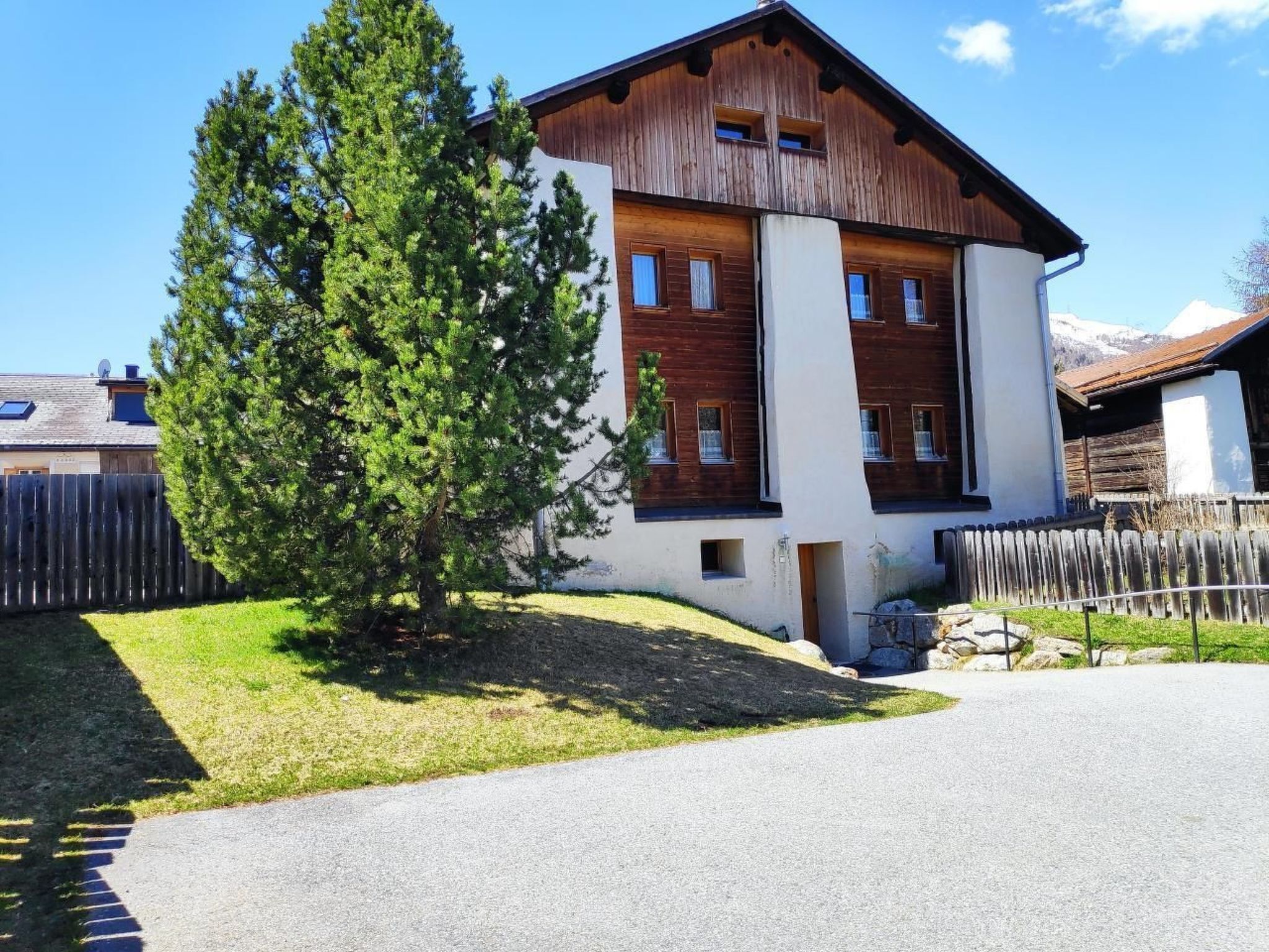 A two-story building with a wooden facade and a white lower section, surrounded by a wooden fence and a large evergreen tree.