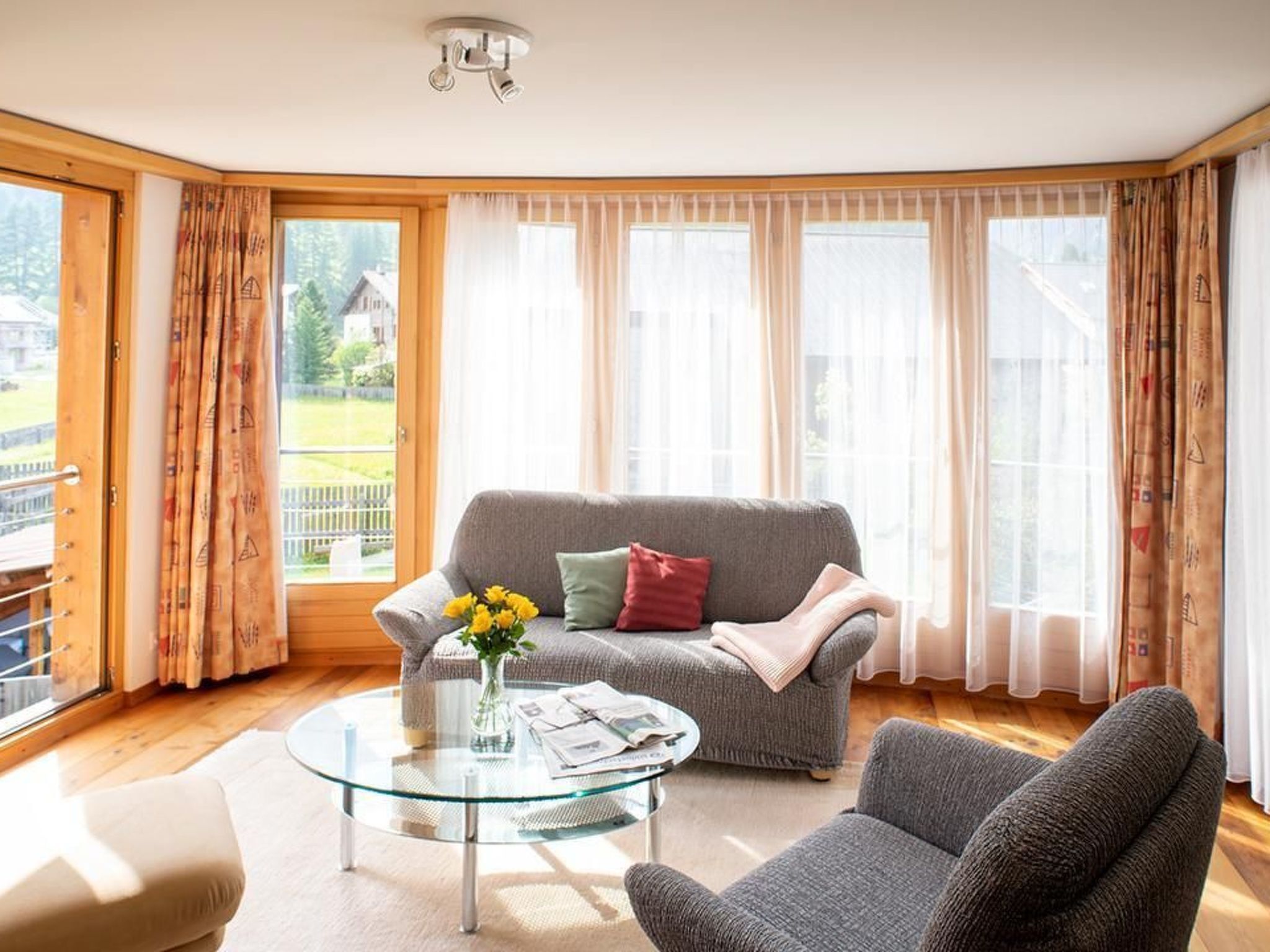 Ein Wohnzimmer mit grauem Sofa, einem Glastisch und Blick auf einen Garten durch große Fenster.