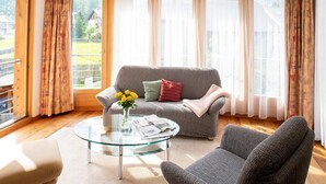 En stue med en grå sofa, et glasbord og udsigt til en have gennem store vinduer.