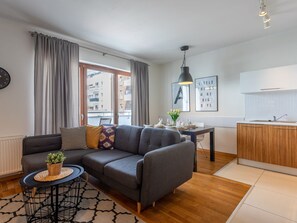 Ein modernes Wohnzimmer mit einem grauen Sofa, einem kleinen runden Tisch mit einer Pflanze und Blick auf die Stadt durch große Fenster.