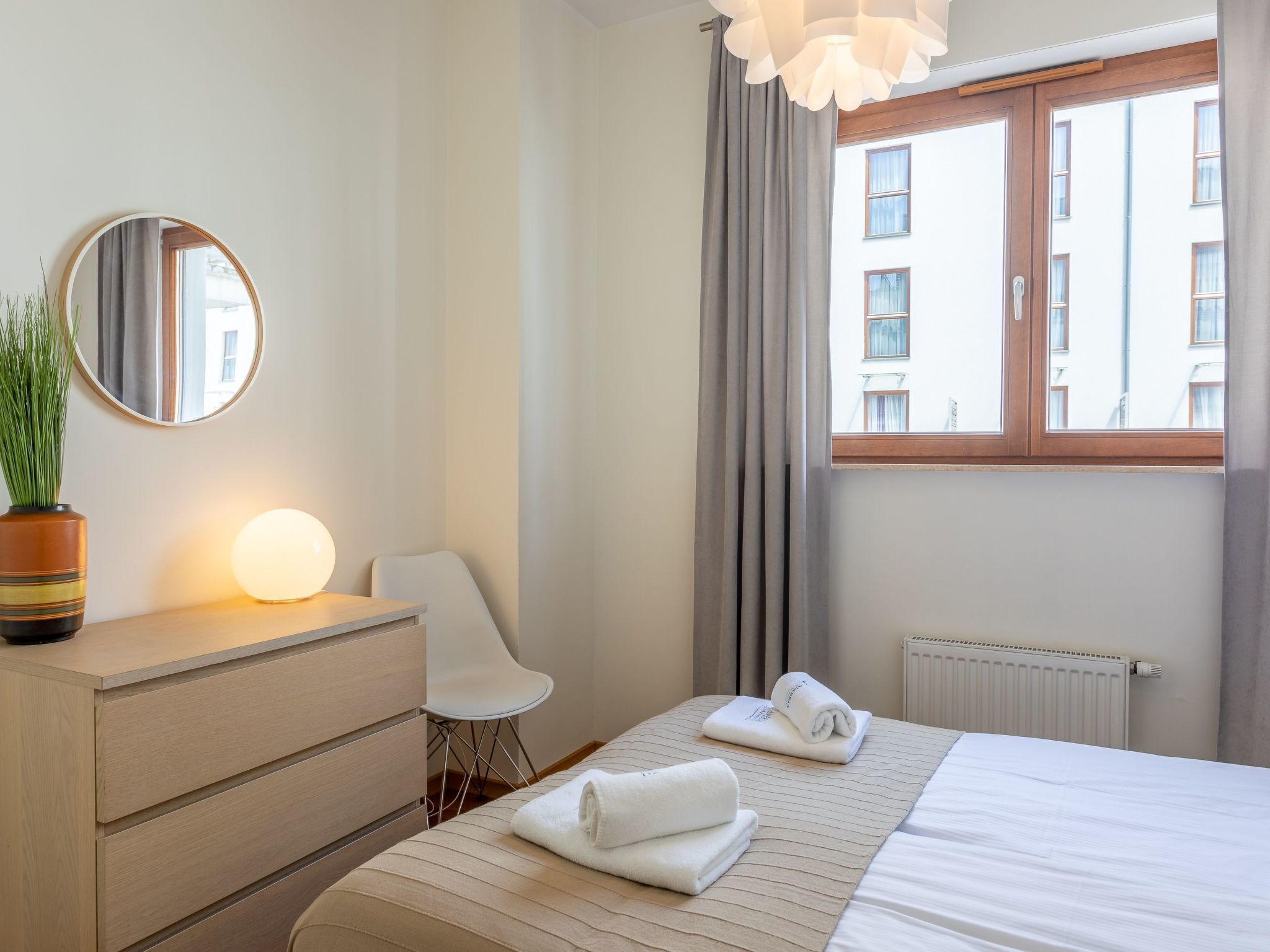 A hotel room with a bed, a bedside table with a lamp, a chair, a potted plant, and a window with curtains.
