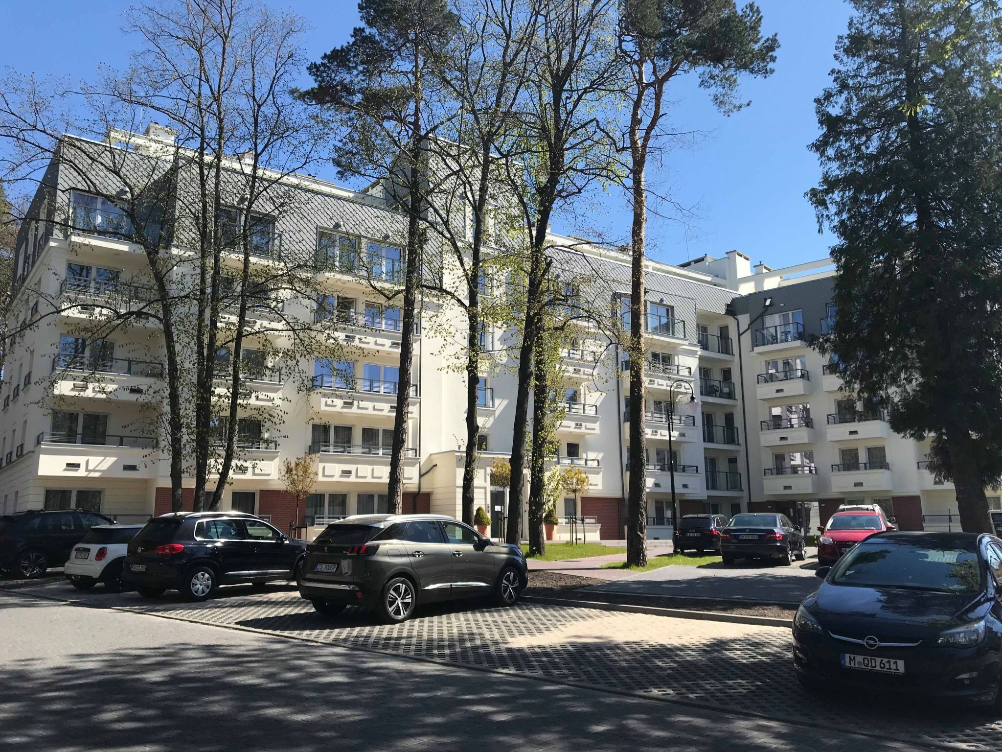 A multi-story residential building with balconies, parked cars, and trees in the foreground.
