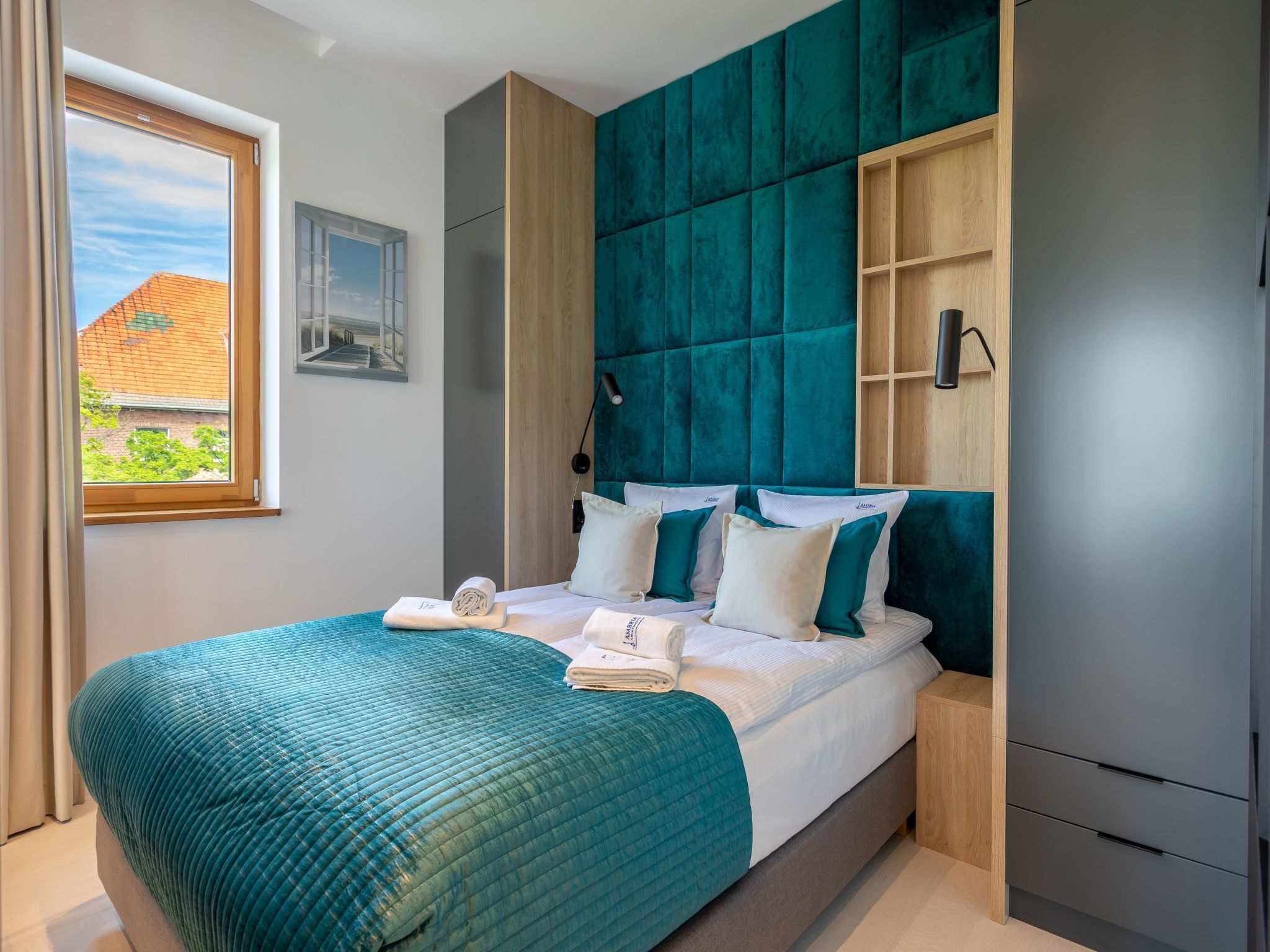 A modern bedroom with a teal upholstered headboard, a wooden nightstand, and a view of a building through a window.
