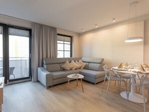 A modern living room with a grey sofa, a small round table, and a dining area with white chairs and a table set for a meal. - Schönes Ferienappartement Gold in Swinemünde by Interhome (Swinoujscie)