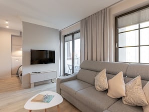 A modern living room with a grey sofa, a flat-screen TV mounted on the wall, a small white side table, and a large window with curtains. - Schönes Ferienappartement Gold in Swinemünde by Interhome (Swinoujscie)