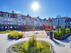 A playground with swings and climbing structures, surrounded by a paved area with greenery and a row of multi-story buildings in the background. - Schönes Ferienappartement Gold in Swinemünde by Interhome (Swinoujscie)