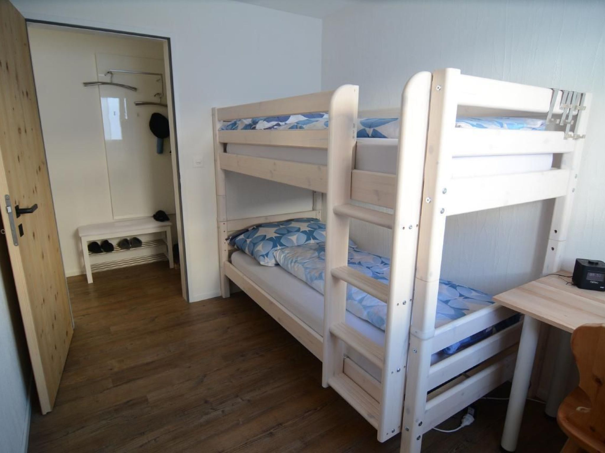 Een slaapkamer met een stapelbed, een houten vloer, een klein bureau en een bank met schoenen.