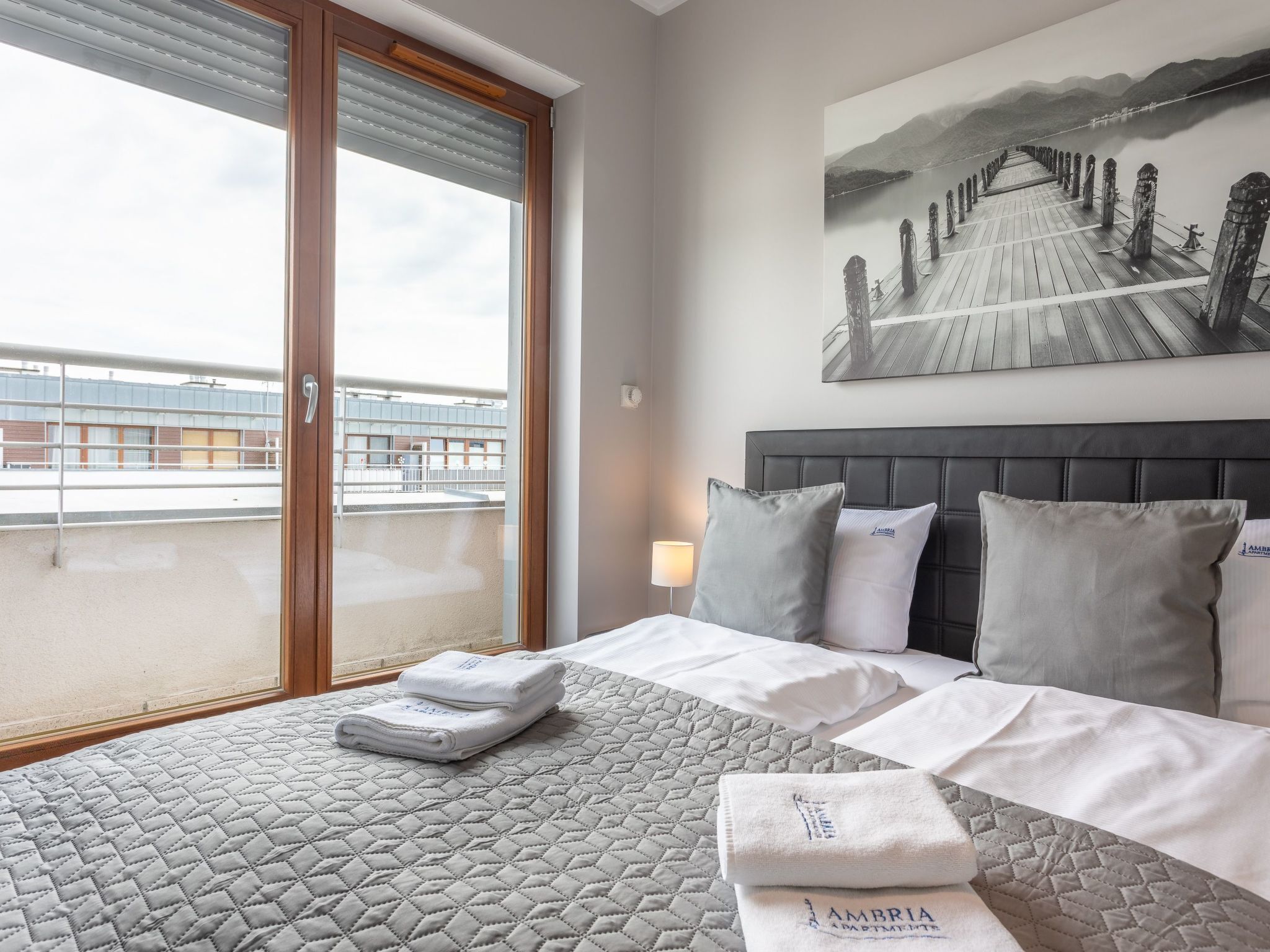 Hotelkamer met een bed, kussens, een gewatteerde spreidek en een raam met jaloezieën, plus een schilderij van een pier.