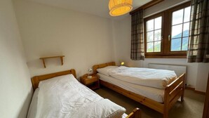 Una habitación pequeña y ordenada con dos camas de madera, una mesita de noche con lámpara, una ventana con cortinas y una lámpara colgante.
