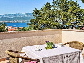Une table recouverte d’une nappe blanche, deux chaises, une tasse et un vase avec des plantes vertes, le tout sur un balcon donnant sur une vue côtière avec des maisons et des arbres.